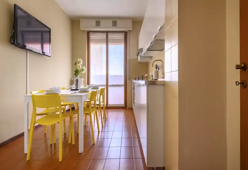Cuisine moderne et lumineuse avec coin repas intégré : table blanche et chaises jaunes, lumière naturelle par une porte vitrée et kitchenette compacte avec placards hauts.