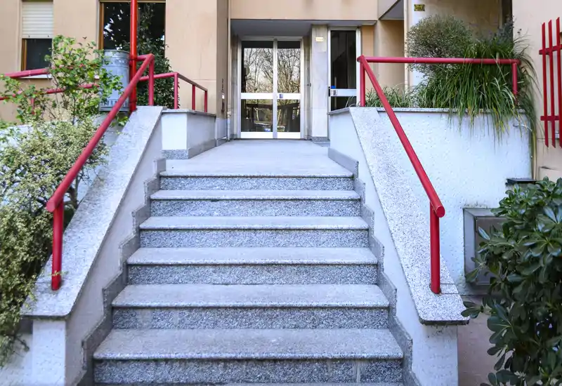 Entrée extérieure avec un large escalier en pierre menant à des portes vitrées ; des mains courantes rouges et des plantations encadrent l'accès.