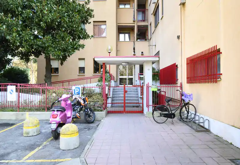 Façade extérieure d'un immeuble montrant l'entrée grillagée, les marches, des scooters et vélos garés, et une cour pavée.