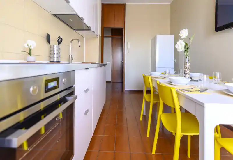 Cuisine moderne et lumineuse avec un long plan de travail, four encastré et une table dressée accompagnée de chaises jaunes vives ; décor floral soigné.