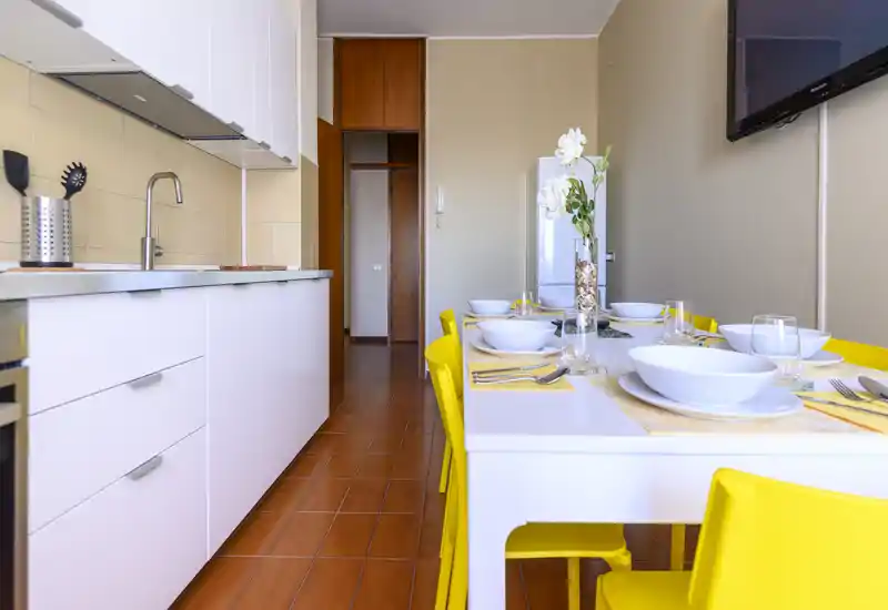 Cuisine moderne et lumineuse avec rangements blancs et table dressée, accompagnée de chaises jaunes vives. L'espace est bien éclairé et mis en scène de façon soignée pour cuisiner et dîner.