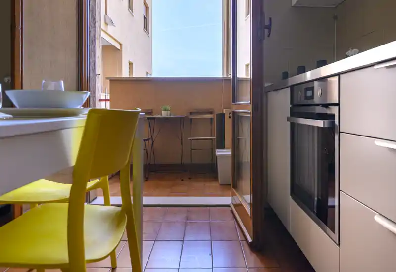 Cuisine moderne compacte s'ouvrant sur un petit balcon ensoleillé. Meubles blancs, four encastré et table avec chaises jaunes apportent une ambiance fraîche et accueillante, baignées de lumière naturelle.