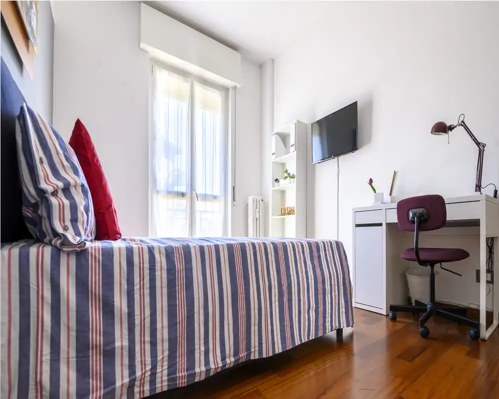 Bright, modern bedroom with a striped single bed, pillows and a small work desk with a chair and wall-mounted TV. Natural light floods the room through a large window, creating a clean and welcoming space ideal for a solo traveler or student.
