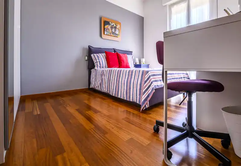 Dormitorio moderno y luminoso con cama a rayas, cojines coloridos y un pequeño espacio de trabajo junto a la ventana — suelo de madera pulida y luz natural suave.