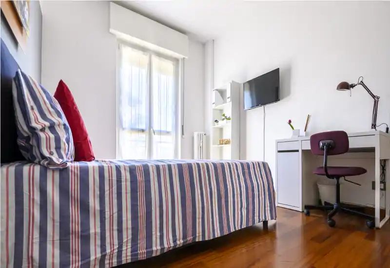 Dormitorio individual luminoso y moderno con cama de rayas, escritorio y TV de pared; gran ventana que aporta abundante luz natural y suelos de madera cálidos.