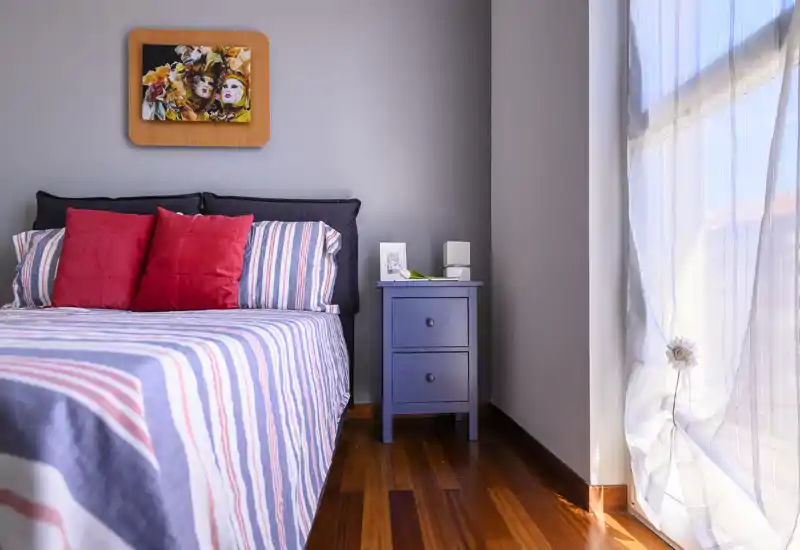 Dormitorio luminoso y bien decorado con colcha a rayas, cojines rojos como acento y una mesita junto a una gran ventana que inunda la habitación de luz natural.