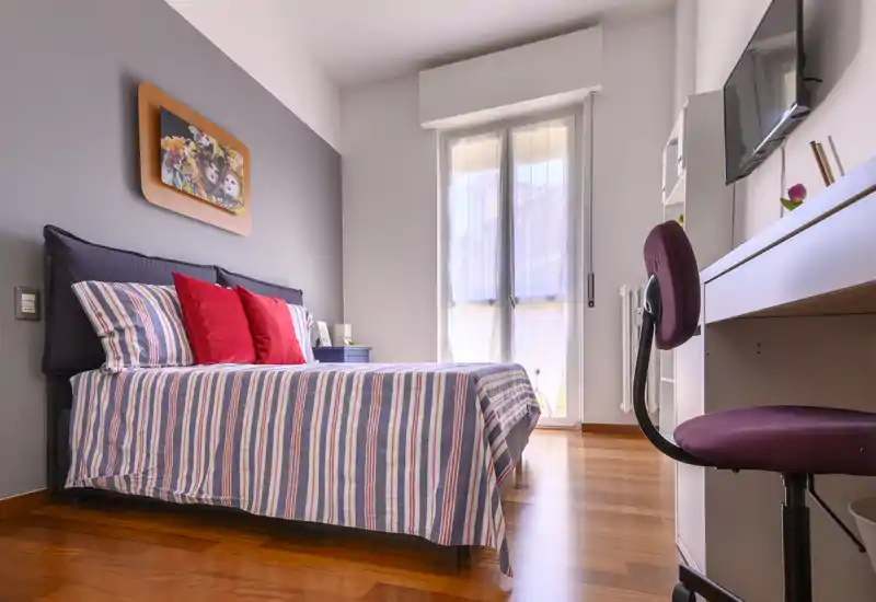 Dormitorio luminoso y bien presentado con cama doble vestida con ropa de rayas y cojines coloridos, suelo de madera y escritorio junto a la ventana — perfecto para una estancia cómoda.