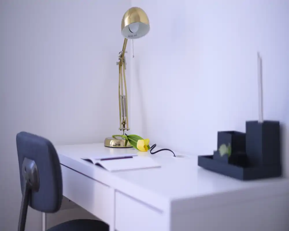 Bright minimal desk corner with a gold adjustable lamp, a single yellow tulip and a chair — a tidy home office or workspace nook.