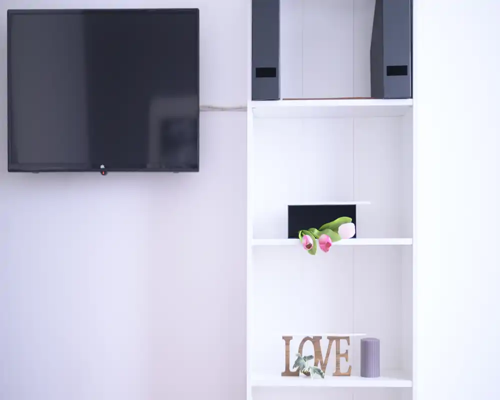 Minimal living area detail with a wall-mounted TV and neat white shelving decorated with flowers and a decorative 'LOVE' sign; bright, calm and suitable for listing photos.