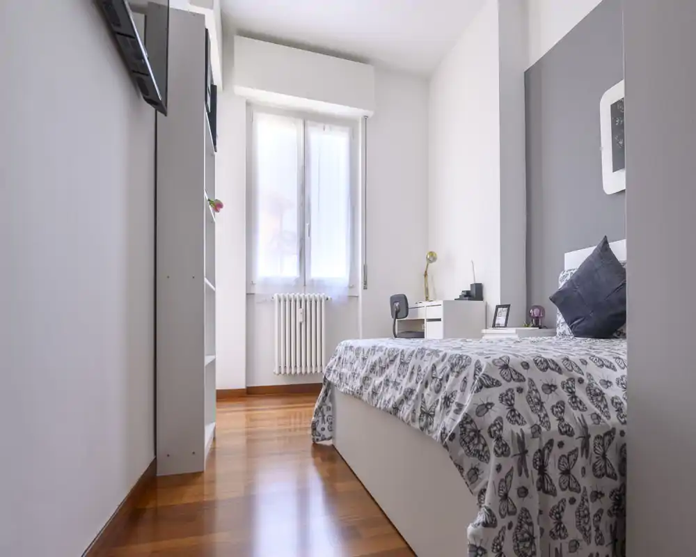 Bright, modern single bedroom with patterned bedding, a study desk by the window and warm wooden floors — well lit and neatly styled for rental listings.