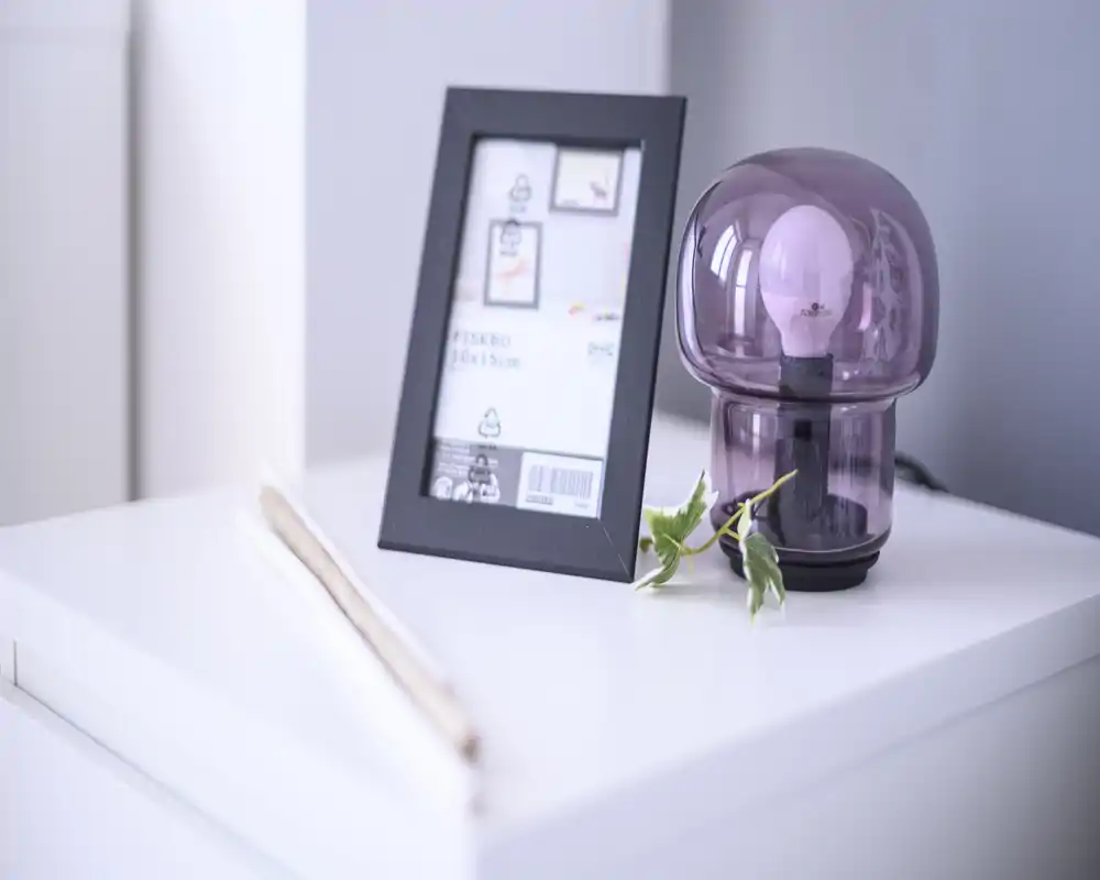 Close-up of a bedside table styled with a modern purple glass lamp, small green plant and a black picture frame on a white nightstand — bright, minimal and contemporary.