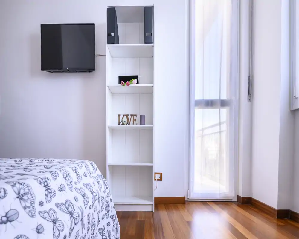 Bright, minimal bedroom with patterned bedding, wall-mounted TV and open shelving beside a tall window letting in ample natural light.