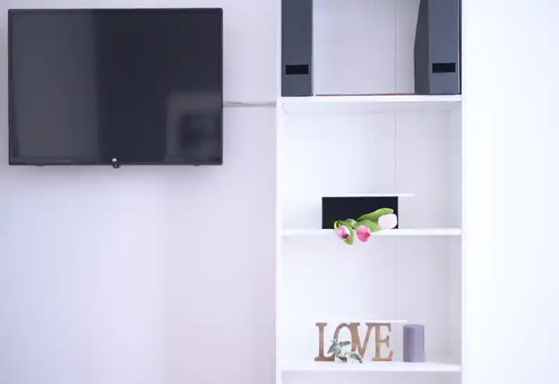 Détail d'un séjour minimaliste avec TV murale et étagères blanches décorées de fleurs et d'un objet décoratif « LOVE » ; clair, calme et adapté aux photos d'annonce.