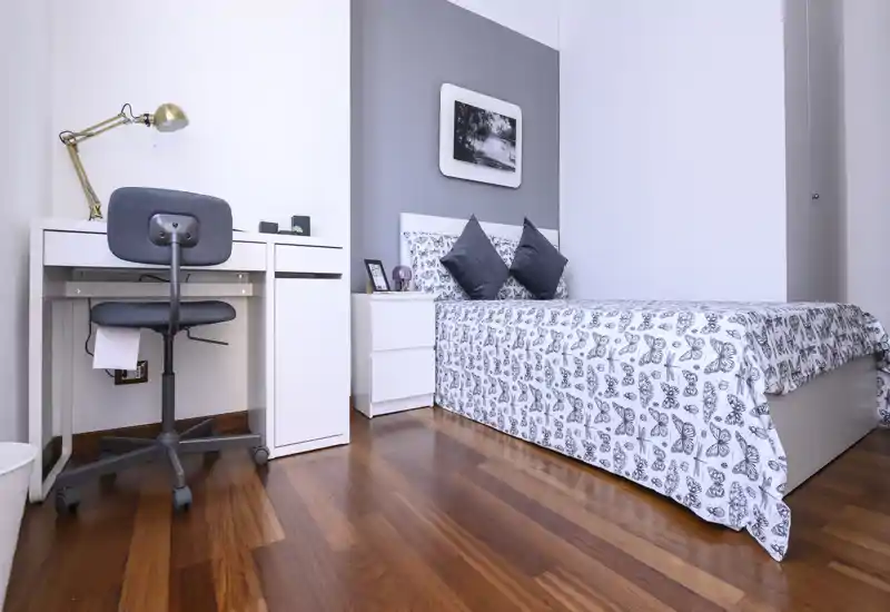 Chambre lumineuse et bien mise en scène avec couette à motif, deux coussins décoratifs et un petit bureau — idéale pour une personne seule ou un étudiant.