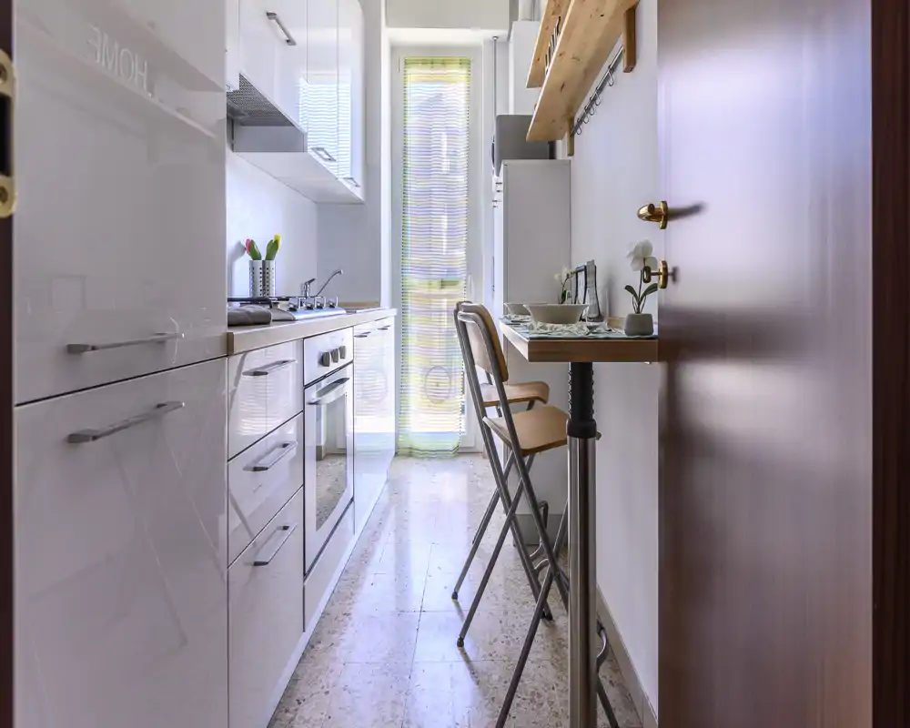 Bright narrow galley kitchen with glossy cabinets, built-in oven and a small breakfast bar with stools by the window — neat, modern and well lit.