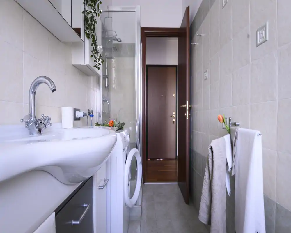 Bright, modern bathroom with a wall-mounted sink, visible shower enclosure and a washing machine; light tiles and neat towels create a clean, functional space.