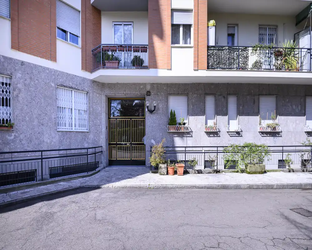 Exterior view of a residential building entrance with a gated doorway, balcony planters and pavement; bright daylight highlights architectural details.
