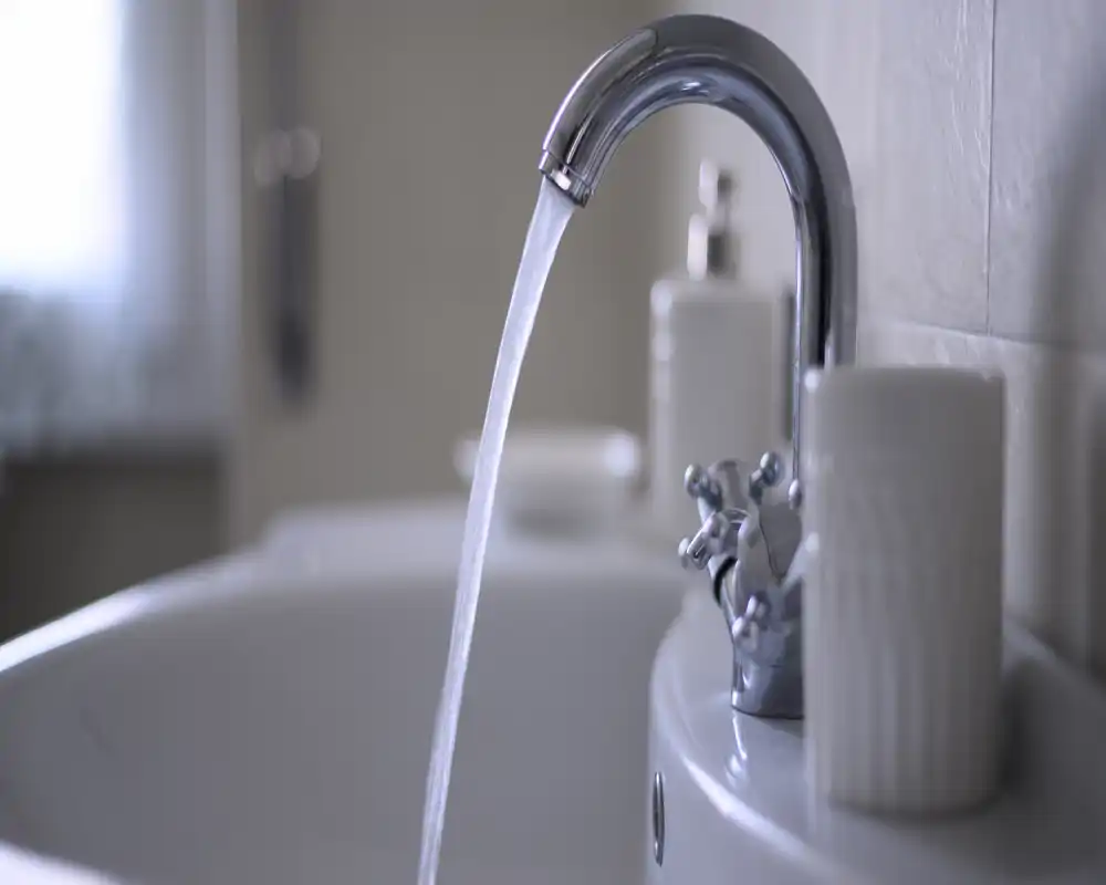 Close-up of a bathroom sink and chrome faucet with a steady stream of water; soft background and neutral tones create a clean, modern feel.