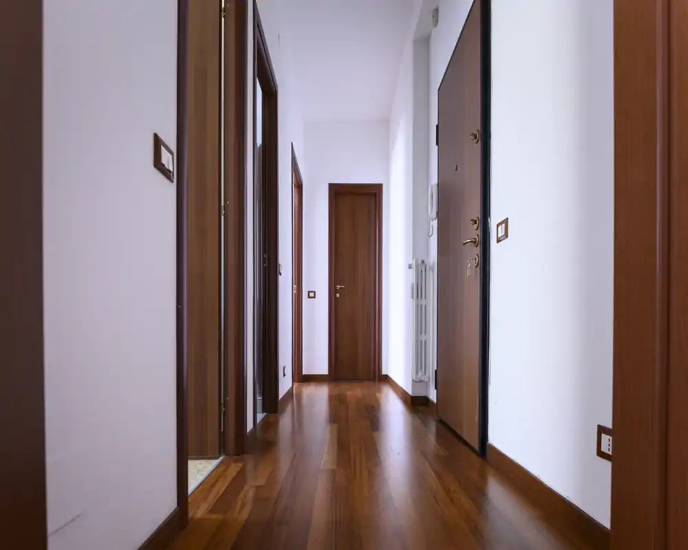 Bright, clean entrance hallway with warm wood flooring, white walls and multiple wooden doors; simple and well-lit space ideal for showcasing an apartment's entry.