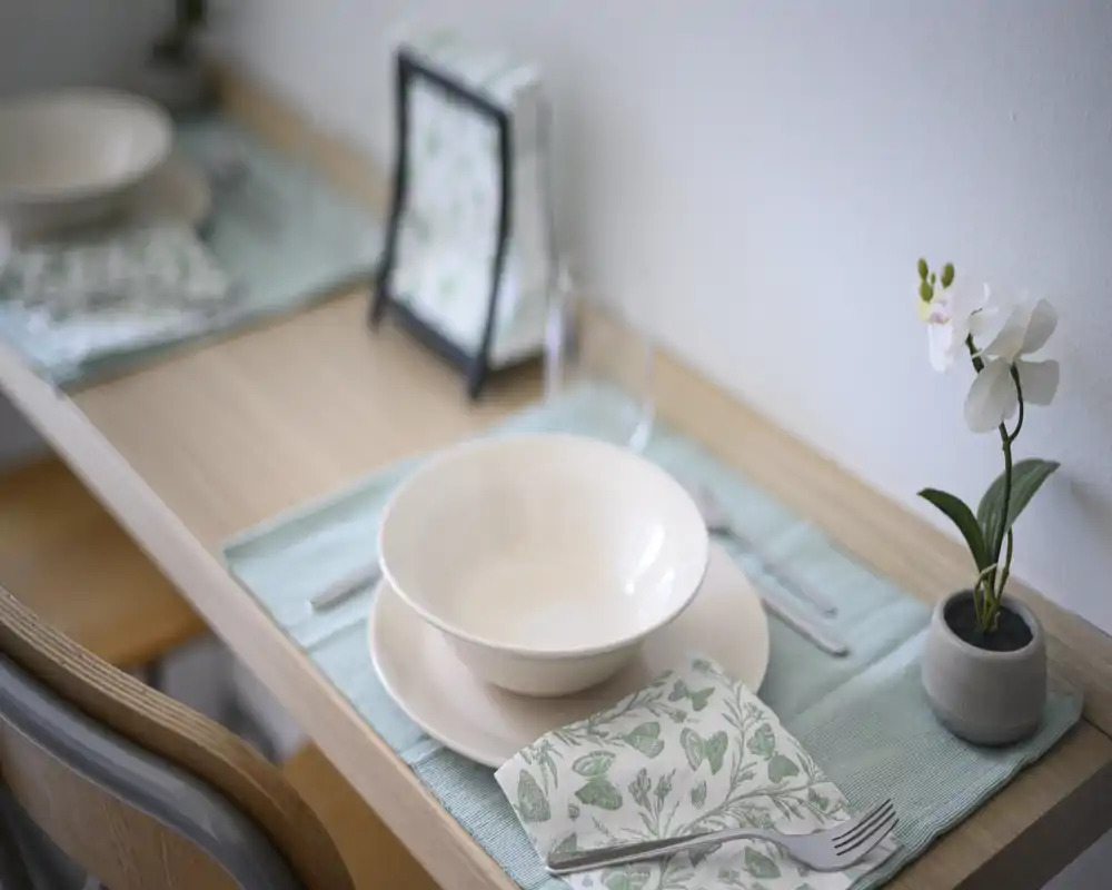 Styled dining nook with a neatly set place: bowl and plate on a pastel placemat, patterned napkin and small orchid for a fresh, minimal table setting ideal for compact apartments.