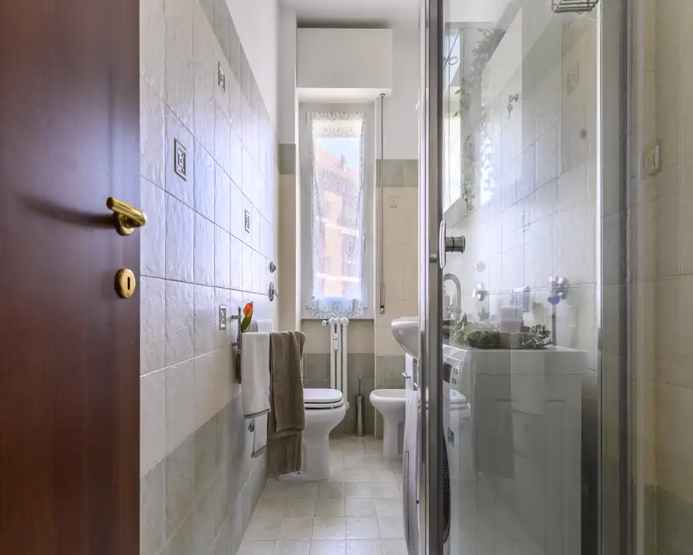 Bright, well-maintained bathroom with a shower cubicle, toilet, bidet and a sink; neutral tiles and a tall window provide natural light.