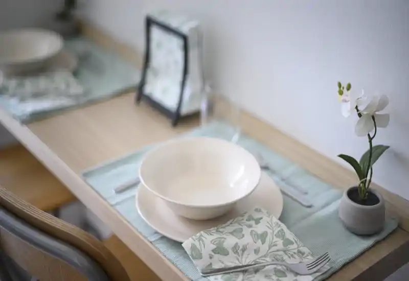 Coin repas soigné avec une table dressée: bol et assiette sur une nappe pastel, serviette à motif et petite orchidée pour une mise en scène fraîche et minimaliste, idéale pour les petits espaces.