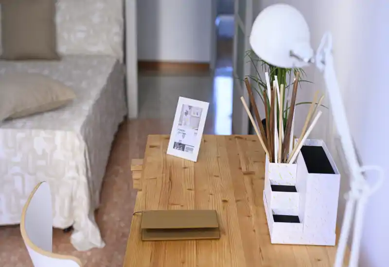 Coin chambre lumineux et chaleureux avec un bureau en bois et un lit partiellement visible drapé de linge neutre; des organiseurs et une lampe offrent un espace de travail accueillant.