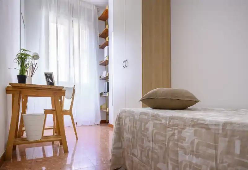 Chambre claire et minimaliste avec un lit simple au premier plan, un bureau en bois près d'une fenêtre aux voilages et une armoire encastrée — un espace soigné et aérien, idéal pour un étudiant ou une personne seule.
