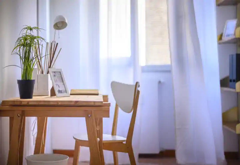 Coin bureau lumineux et minimaliste avec bureau et chaise en bois près d'une fenêtre aux voilages doux ; plante et photo encadrée apportent une touche chaleureuse.