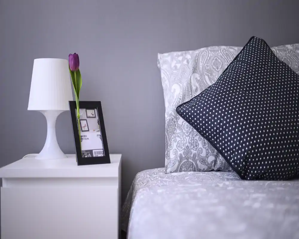 Coin de chambre lumineux et chaleureux avec une literie à motifs, un coussin décoratif à pois et une table de chevet blanche portant une lampe et une tulipe violette.