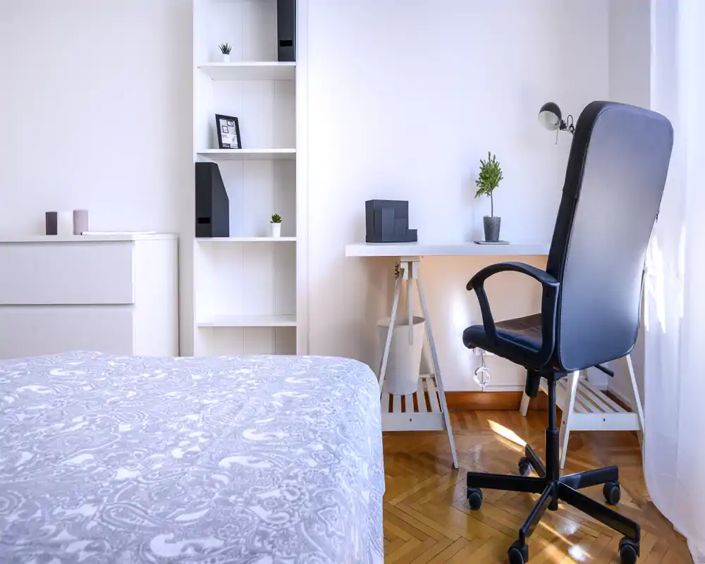 Chambre lumineuse et minimaliste avec une couette à motifs, des étagères blanches et un bureau avec chaise pivotante près d'une fenêtre ensoleillée.