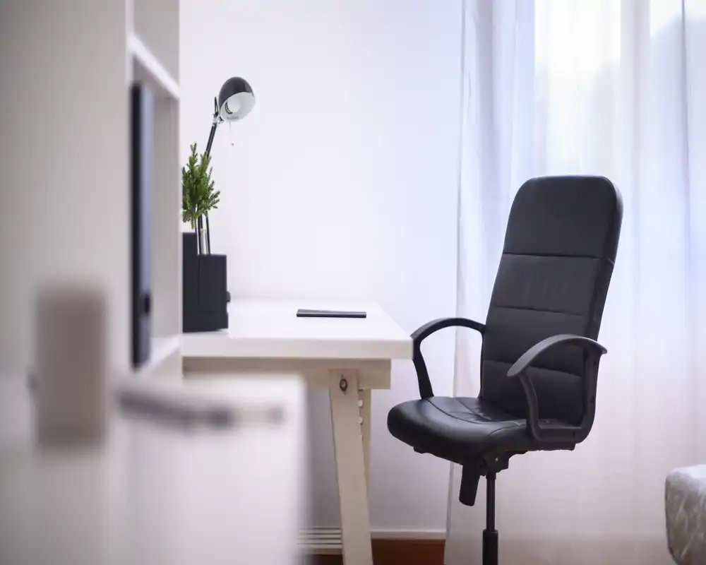 Coin bureau lumineux et minimaliste avec un bureau blanc, une chaise pivotante noire, une lampe de bureau et une petite plante près d'une fenêtre à voilage.