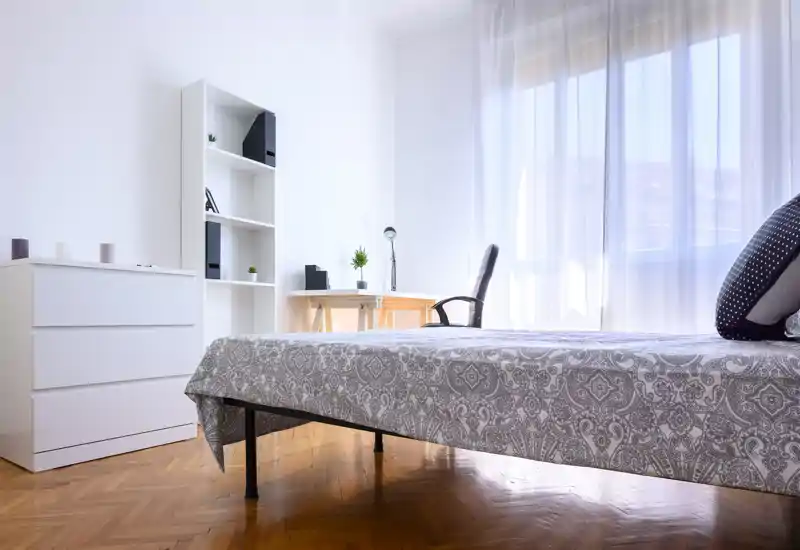 Chambre lumineuse et minimaliste avec une couette à motif, grande fenêtre avec voilages et un petit bureau — espace propre, aéré et bien éclairé.