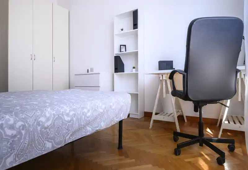 Chambre lumineuse et minimaliste avec couvre-lit à motif, rangements blancs et un espace bureau avec chaise de bureau près d'une fenêtre ensoleillée.