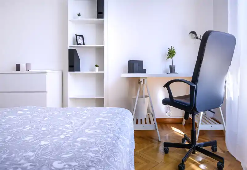 Chambre lumineuse et minimaliste avec une couette à motifs, des étagères blanches et un bureau avec chaise pivotante près d'une fenêtre ensoleillée.