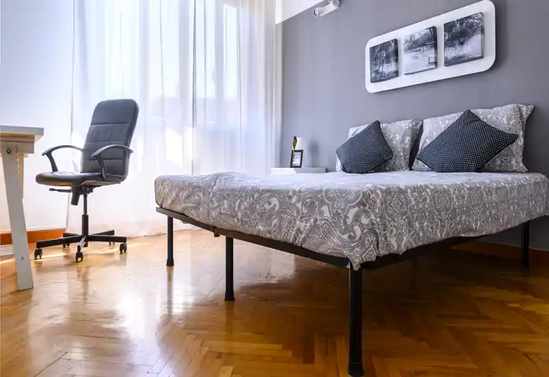 Chambre moderne et lumineuse avec lit double à motif, coussins décoratifs et lumière naturelle provenant d'une grande fenêtre ; parquet et chaise de bureau.
