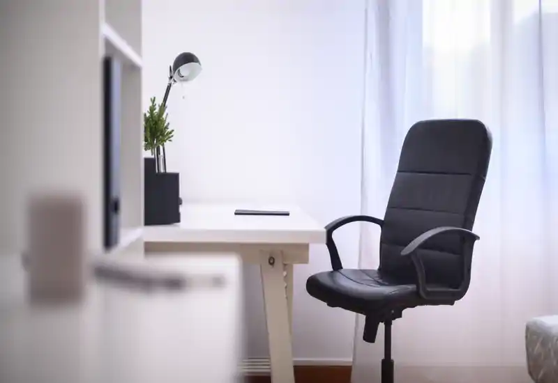 Angolo lavoro luminoso e minimal con scrivania bianca, lampada da tavolo, piccola pianta e sedia ufficio nera vicino alla finestra con tenda leggera. Pulito, moderno e ben illuminato, ideale per smart working.