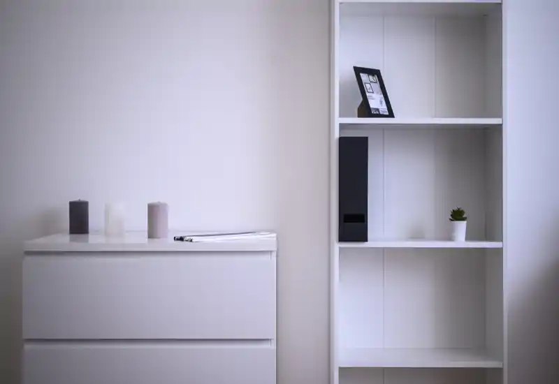 Angolo di camera minimale con comò bianco e libreria adiacente, decorato con candele, una foto incorniciata e una piantina; pulito, luminoso e minimal.