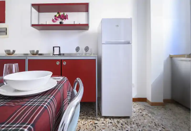 Angolo cucina compatto e curato con pensili rossi, frigorifero bianco alto e tavolo apparecchiato – luminoso e accogliente per pasti informali.