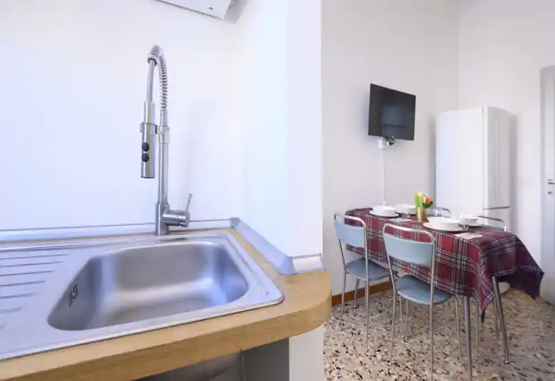 Angolo cucina luminoso e compatto con lavello in acciaio inox in primo piano e un piccolo tavolo da pranzo con tovaglia a quadri rossa, sedie e frigorifero sullo sfondo.