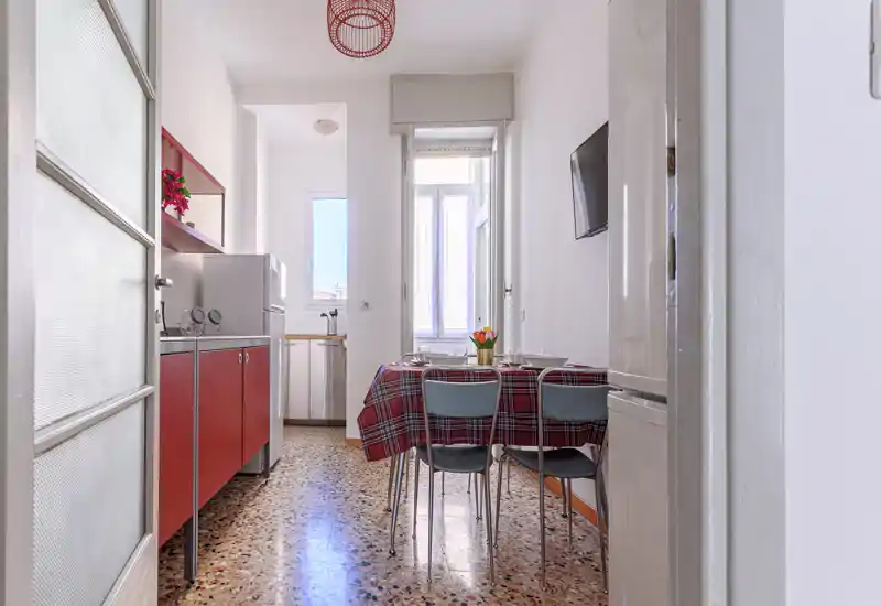 Cucina compatta e luminosa con mobili rossi, un piccolo tavolo per quattro persone e luce naturale dalla finestra — perfetta per pasti informali.