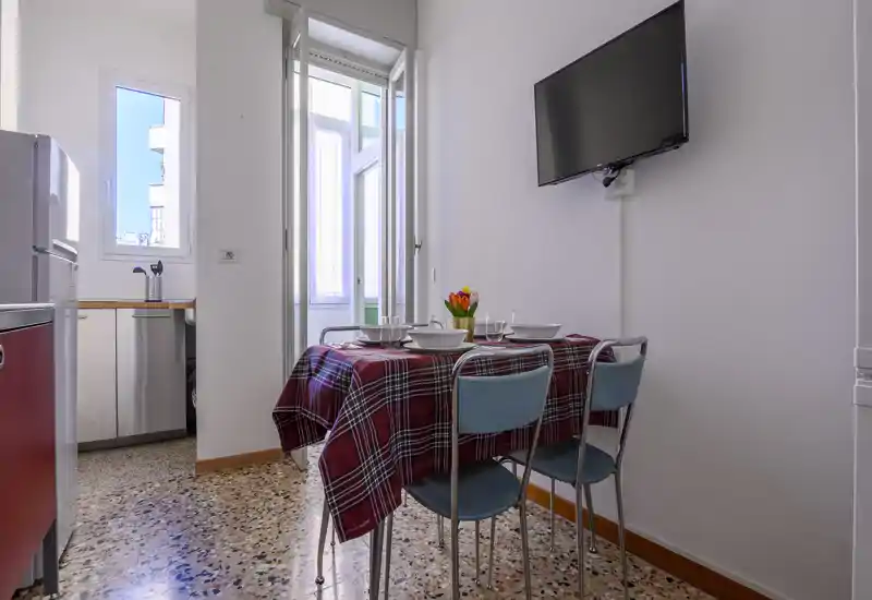 Angolo pranzo compatto con tavolo apparecchiato, tovaglia tartan rossa, quattro sedie e luce naturale proveniente dalla finestra e dalla porta adiacente.