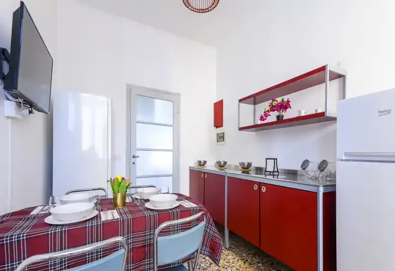 Cucina compatta e luminosa con mobili rossi, tavolo apparecchiato per quattro e elettrodomestici moderni — ideale per pasti accoglienti in uno spazio di stile.