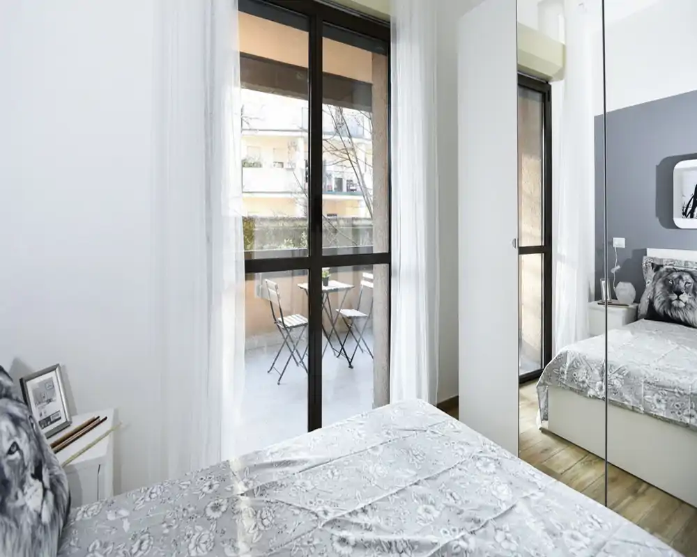 Dormitorio moderno y luminoso con cama doble, textiles decorativos y puerta de cristal que da a un pequeño balcón con mesa. Estilo limpio, armario con espejo y mucha luz natural que hacen la habitación amplia y acogedora.