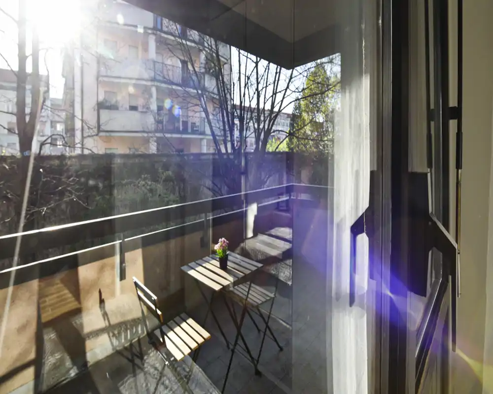 Balcón privado soleado con pequeña mesa bistró y dos sillas, ideal para el café de la mañana; puerta de cristal con vistas a árboles y edificios cercanos.