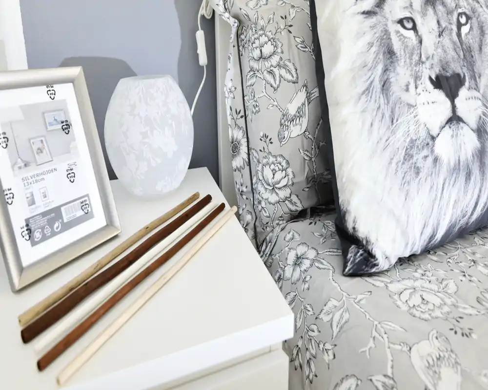 Detalle recortado de dormitorio que muestra una mesita de noche con objetos decorativos y una porción de la cama con ropa de cama estampada y un cojín con león.