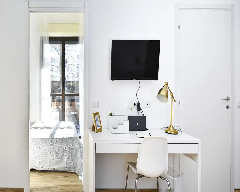 Vista luminosa y moderna de un dormitorio a través de una puerta, con la cama bien hecha y colcha estampada; en primer plano un escritorio blanco, silla y TV de pared, ideal para un piso compacto.