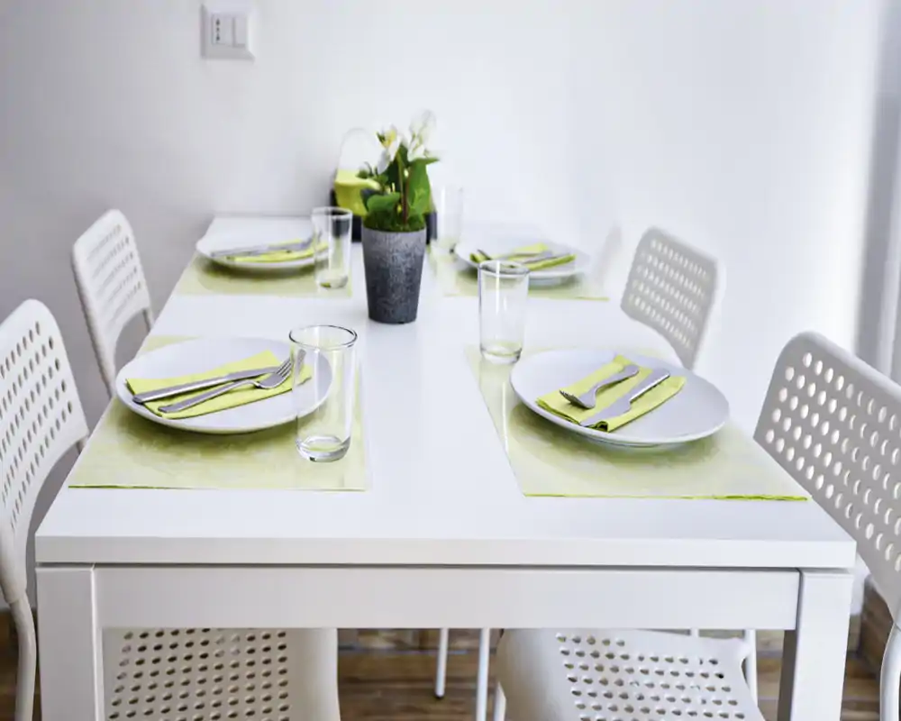 Mesa de comedor blanca moderna preparada para cuatro con platos sencillos, cubiertos y una pequeña planta como centro; luminosa, minimalista y lista para una comida informal.