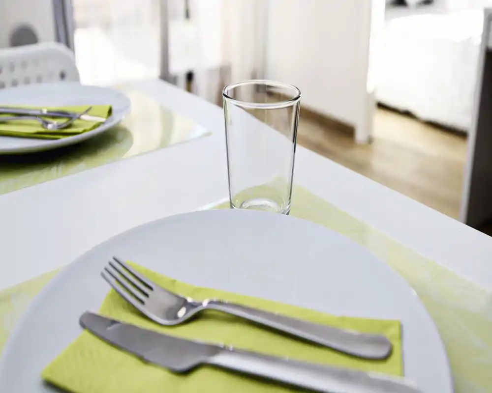 Plano detalle de una mesa puesta con plato, cubiertos y vaso sobre un salvamantel claro; luz natural y presentación ordenada que hacen el rincón de comedor acogedor.