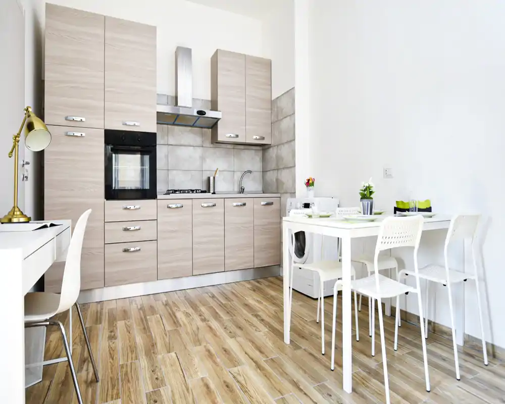 Cocina tipo kitchenette luminosa con muebles de madera clara, salpicadero de azulejos y mesa de comedor compacta para cuatro — limpia, bien iluminada y funcional.